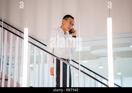 Un homme chinois asiatique dans une chemise en chemise et pantalon professionnel walking down steps pendant qu'il parle sur son smartphone. Banque D'Images