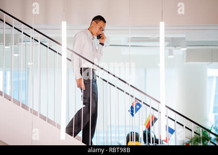 Un homme chinois asiatique dans une chemise en chemise et pantalon professionnel walking down steps pendant qu'il parle sur son smartphone. Banque D'Images