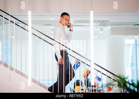 Un homme chinois asiatique dans une chemise en chemise et pantalon professionnel walking down steps pendant qu'il parle sur son smartphone. Banque D'Images