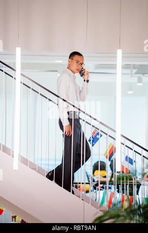 Un homme chinois asiatique dans une chemise en chemise et pantalon professionnel walking down steps pendant qu'il parle sur son smartphone. Banque D'Images