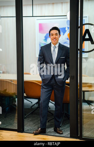 Portrait of a handsome, sains et sûrs d'affaires asiatiques en costume et cravate debout dans une salle de réunion. Il sourit alors qu'il s'appuie sur une table. Banque D'Images