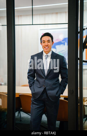 Portrait of a handsome, sains et sûrs d'affaires asiatiques en costume et cravate debout dans une salle de réunion. Il sourit alors qu'il s'appuie sur une table. Banque D'Images