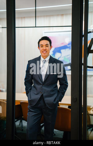 Portrait of a handsome, sains et sûrs d'affaires asiatiques en costume et cravate debout dans une salle de réunion. Il sourit alors qu'il s'appuie sur une table. Banque D'Images