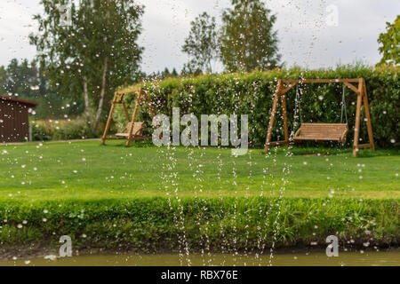 Deux bancs balançoire en bois dans le jardin avec des gouttes d'eau au premier plan. Fontaine eau gouttes dans l'air avec des balançoires de jardin à l'arrière-plan Banque D'Images