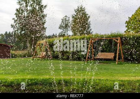 Deux bancs balançoire en bois dans le jardin avec des gouttes d'eau au premier plan. Fontaine eau gouttes dans l'air avec des balançoires de jardin à l'arrière-plan Banque D'Images