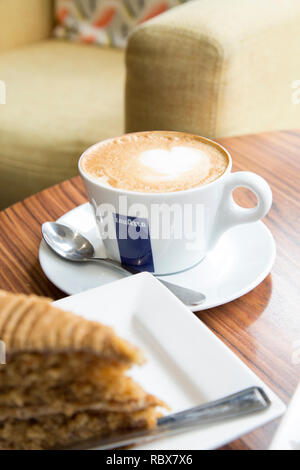 Tasse de cappuccino Lavazza café sur une soucoupe avec une cuillère et une tranche de gâteau au premier plan Banque D'Images