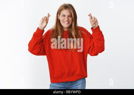 Optimiste et charismatique jeune femme en rouge pull surdimensionné élever les mains avec les doigts croisés pour la bonne chance sourire sentiment fortune et chance de son côté, foi dans WIN et succès Banque D'Images