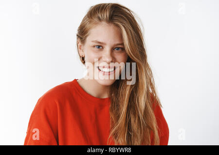 Heureuse et sans soucis sincère charmante jeune femme charismatique aux yeux bleus, cheveux blonds et freckels tout sourire avec un sourire parfait, Porter du rouge pull-over chaud, posant contre mur gris Banque D'Images