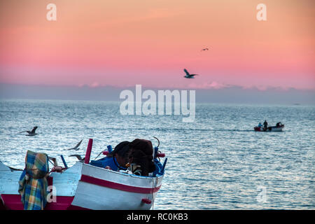 Vol de mouettes au-dessus de bateaux échoués sur la plage avec un arrière-plan spectaculaire coucher du soleil, à Oued Lao, un petit village de pêcheurs sur la côte de la province Banque D'Images