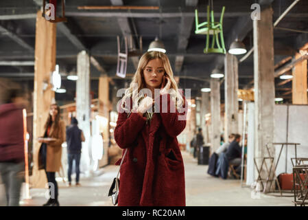 Fashion femme occupée à la rue du marché. Jeune femme blonde en rouge élégant manteau de fourrure eco parle par smartphone à street market Banque D'Images