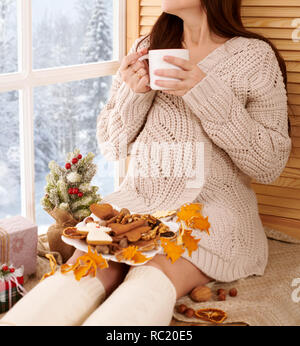 Femme enceinte assis près de la fenêtre et de boire le thé, le magnifique paysage hivernal avec la forêt enneigée est en dehors de la fenêtre, décoration de Noël Banque D'Images