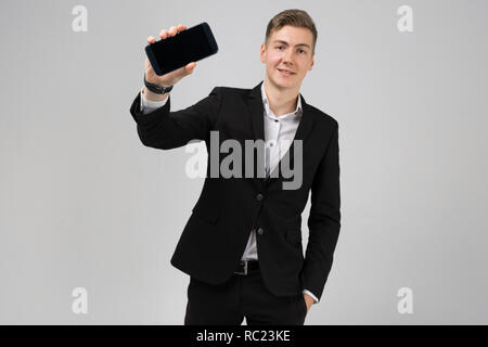 Portrait de jeune homme en costume noir montre téléphone portable avec écran blanc isolé sur fond blanc Banque D'Images