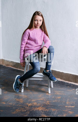 Confiant enfant girl fashion posent sur chaise,fond blanc en béton. Modèle élégant en rose pull. Beau gosse glamour adolescent,u,occasionnels Banque D'Images