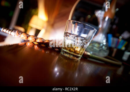 Incliné verre de whisky sur le bar avec un narguilé sur un arrière-plan flou. Banque D'Images