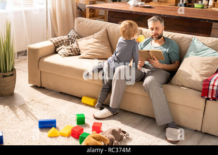 Beau graves père barbu assis sur un canapé confortable avec des oreillers et de la lecture livre alors que petit fils père demander à jouer jeu actif Banque D'Images