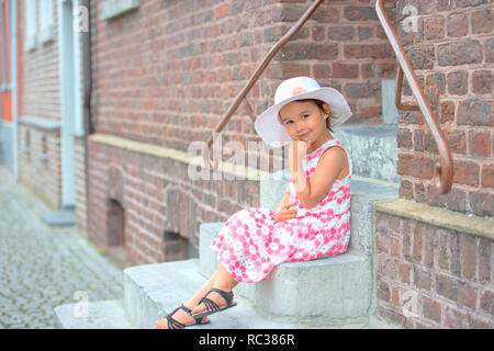 Adorable petite fille wearing white hat sitting on stairs sur jour d'été chaud et ensoleillé Banque D'Images