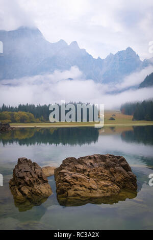 Vue sur le lac de Laghi di Fusine sur un matin brumeux avec quelques rochers à la plage et montagne gamme Schloss Weikersdorf près de Tarvisio en Italie Banque D'Images