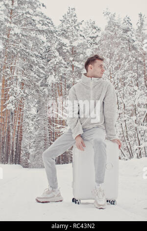 Portrait de jeune homme élégant dans des vêtements blancs marcher sur la neige holding suitcase et rendez pathway forêt de pins. Concept de voyage de vacances d'hiver. Pis Banque D'Images