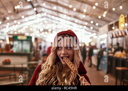 La culture de l'alimentation de rue. Jeune femme blonde à tête rouge et un manteau de fourrure eco eatting saucisse vegan dans la pâte à foire de rue. Banque D'Images