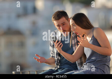 Surpris couple checking smart phone content assis sur une corniche dans une ville Banque D'Images