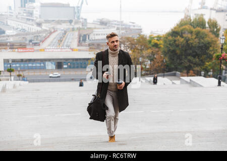 Belle Veste homme portant l'étage à pied à l'extérieur, holding cup de café à emporter, à l'aide de mobile phone Banque D'Images