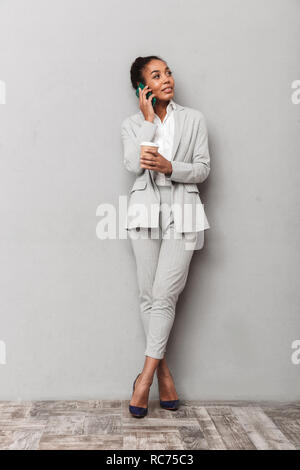 Portrait d'une femme d'affaires de l'Afrique de l'assurance portant sur l'article costume holding fond gris, tasse de café, talking on mobile phone Banque D'Images