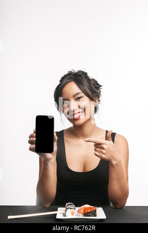 Smiling young asian woman eating sushi à la table isolated over white background, montrant écran blanc téléphone mobile Banque D'Images