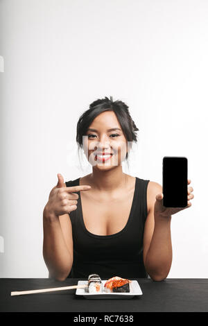 Smiling young asian woman eating sushi à la table isolated over white background, montrant écran blanc téléphone mobile Banque D'Images