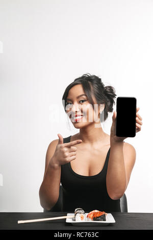 Smiling young asian woman eating sushi à la table isolated over white background, montrant écran blanc téléphone mobile Banque D'Images