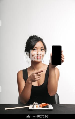 Smiling young asian woman eating sushi à la table isolated over white background, montrant écran blanc téléphone mobile Banque D'Images