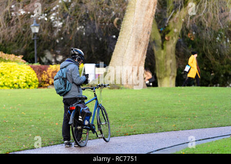 Casque de cycliste de mâle est l'obéissance à l'intérieur du parc et a démonté, debout avec bike & looking at phone - Musée Jardins, York, Yorkshire, Angleterre, Royaume-Uni. Banque D'Images