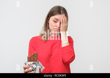 Jeune femme triste fatigué de restrictions de régime holding le chocolat sucré isolé sur fond de mur gris. Concept de la nutrition. Les sentiments de culpabilité Banque D'Images