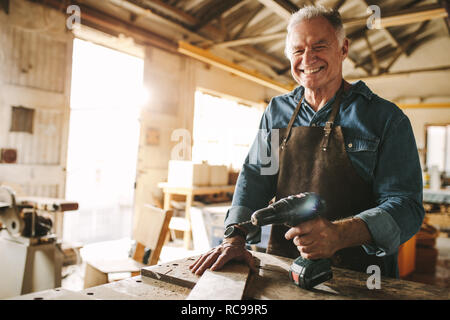 Smiling senior carpenter debout à l'établi avec une perceuse. Mature male carpenter dans son atelier. Banque D'Images