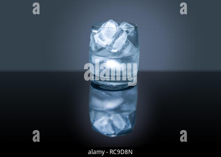 Verre avec des glaçons et de l'eau reflétant sur table avec fond sombre Banque D'Images