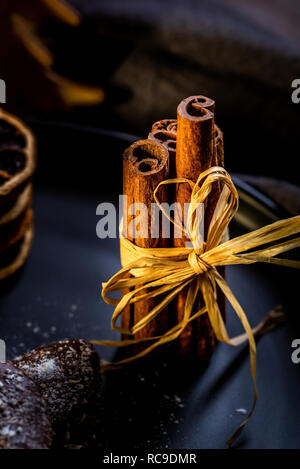 Photo verticale avec plusieurs bâtons de cannelle brun collée par cordon naturel. Bâtonnets sont sur la plaque noire avec des bonbons au chocolat et empilés anneaux orange à sec Banque D'Images