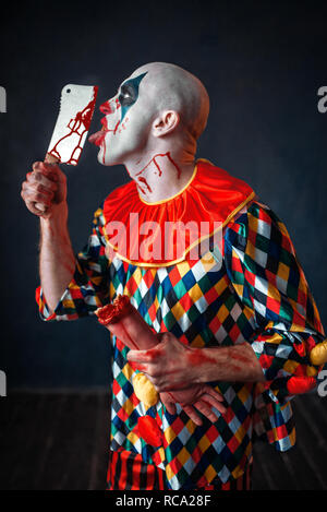 Scary clown sanglant lèche la lame de couteau. Homme avec un miroir en costume halloween, crazy maniac détient main humaine Banque D'Images