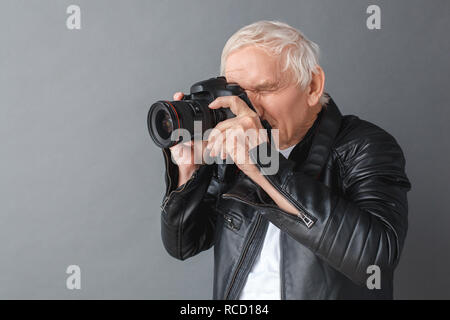 Hauts homme portant veste en cuir noire isolés sur mur gris en prenant des photos avec son appareil photo, vue côté concentré Banque D'Images