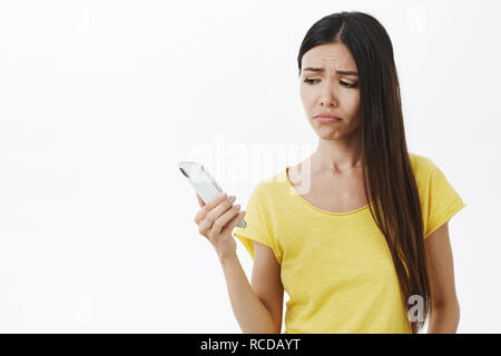 Taille plan sur mécontents en colère belle jeune femme sans maquillage en jaune t-shirt holding smartphone à écran portable à froncer avec sourire sombre triste et solitaire sur fond gris Banque D'Images