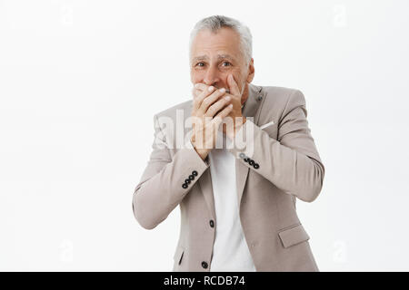 Portrait d'emotive beau vieil homme en costume élégant couvrant la bouche avec palms ne moquant drôle d'ami se sentir désolé et amusé en même temps posant en costume sur fond gris Banque D'Images