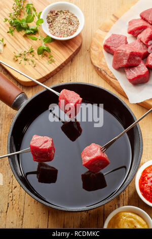 Trois fourchettes à fondue avec des cubes de viande crue suspendu au-dessus d'une casserole d'huile chaude vu du dessus avec des trempettes et des épices qui l'entourent Banque D'Images