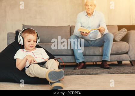 Fossé entre les générations. Adorable petit garçon couché sur le Pouf poire et jouant sur le comprimé dans un casque, tandis que son grand-père assis sur le canapé et la lecture d'un b Banque D'Images