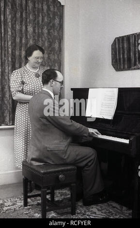 1950s, historique, à la maison une femme debout regardant son mari jouer le piano, Angleterre, Royaume-Uni. À l'époque avant la télévision, le divertissement à domicile était fourni par la radio, les livres et la lecture d'un instrument de musique. Banque D'Images