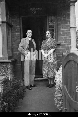 Années 1950, historique, un fier mari et femme habillée dans les vêtements de l'époque dans l'article sur l'extérieur pose officielle path à l'entrée de leur maison, England, UK. Banque D'Images