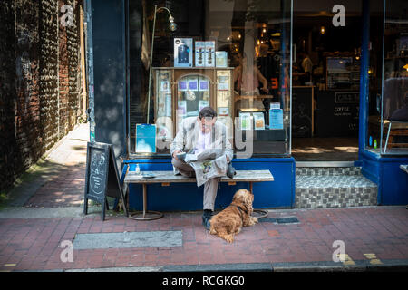 Un homme boit du café et lit un journal avec un chien à ses pieds en restant assis à l'extérieur d'un café à Brighton, East Sussex, Angleterre. Banque D'Images