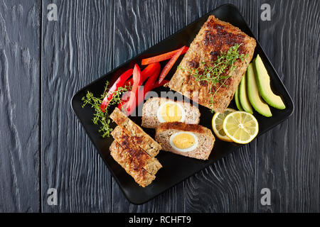 Le pain de viande avec oeuf dur dragées servi avec des tranches d'avocat et de poivron rouge sur un plateau rectangulaire noir sur une table de bois, vue de dessus, Fla. Banque D'Images