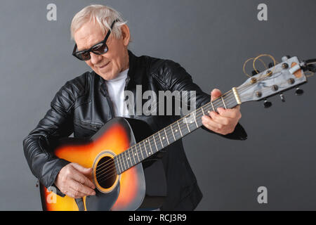 Hauts homme portant veste en cuir noire et lunettes isolés sur mur gris à la guitare qu'à la joyeuse smiling close-up Banque D'Images