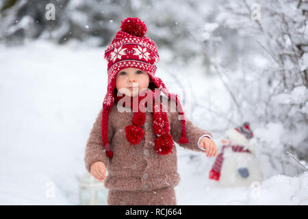 Portrait of a cute toddler bébé vêtu d'un tricot main marron veste, pantalon, bonnet et écharpe rouge, tenant une lanterne et bonhomme de neige, promenades à travers les cimes enneigées Banque D'Images