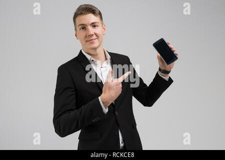 Portrait de jeune homme en costume noir montrant doigt sur téléphone mobile avec écran blanc isolé sur fond blanc Banque D'Images