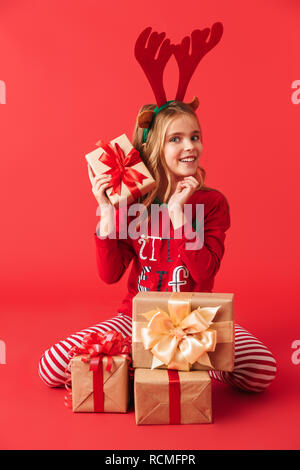 Jolie petite fille portant des cornes de cerf Noël isolé sur fond rouge assis, holding pile de boîtes actuelles Banque D'Images