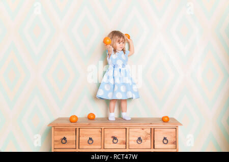 Tout-petit enfant fille en robe bleue debout sur une commode en bois et holding fresh orange mandarine dans ses mains et va les jeter sur fond de losange. Vacances d'hiver ou de noël Banque D'Images